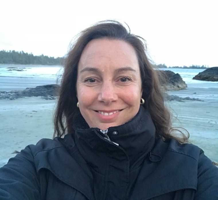 Jennifer smiling on a beach among tidal rocks, wearing a black jacket